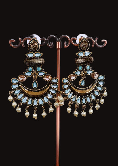 A pair of ornate gold-toned dangle earrings with light blue teardrop stones, a crescent moon design, and dangling pearl beads, displayed on a decorative stand against a light gray background.