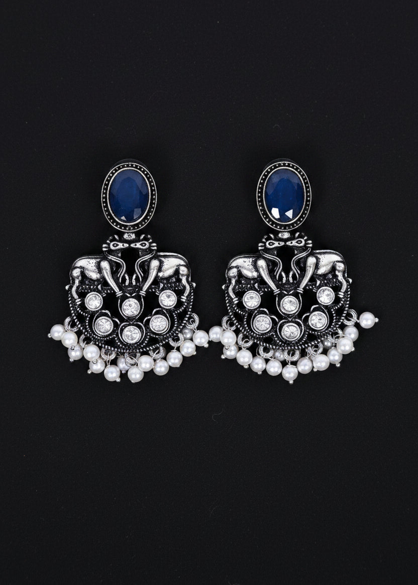 A pair of ornate silver-toned jhumka earrings with oval dark blue stones and dangling pearl-like beads