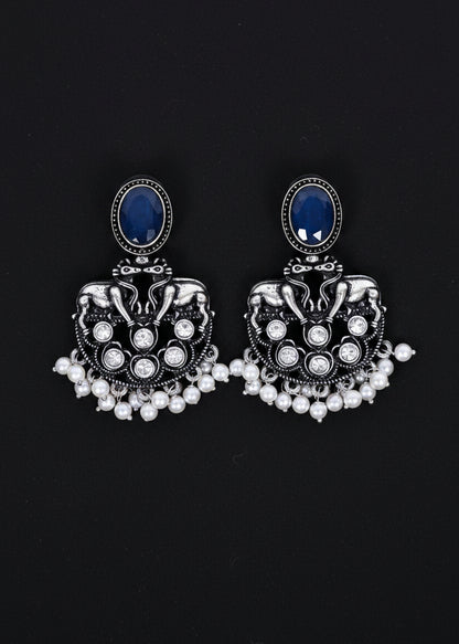 A pair of ornate silver-toned jhumka earrings with oval dark blue stones and dangling pearl-like beads