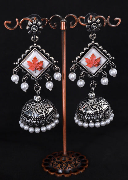 A pair of silver-toned traditional Indian jhumka earrings with white pearl bead drops and a central white square inlay featuring a red maple leaf.