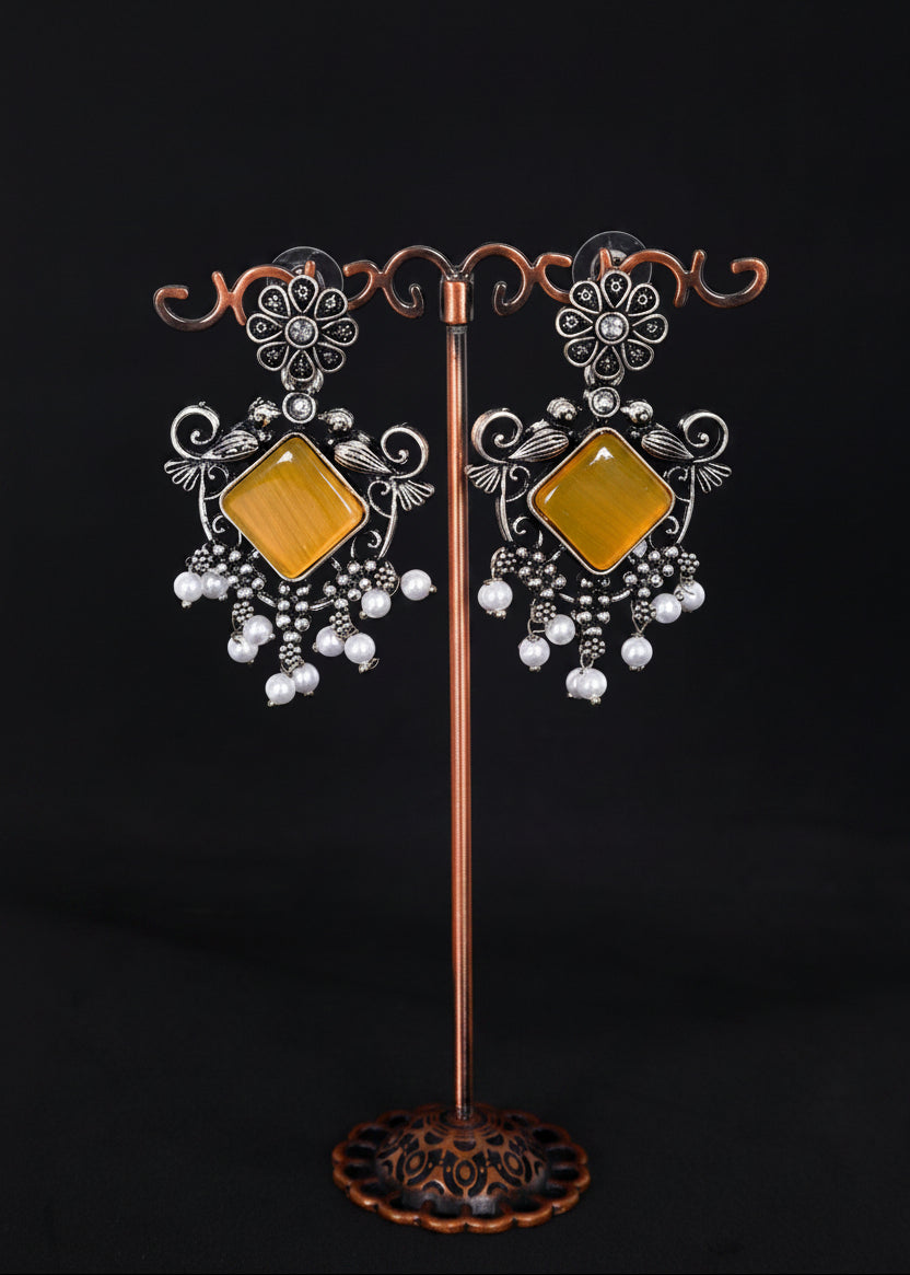 A pair of ornate silver-toned statement earrings with large square amber-yellow centers and dangling white pearl-like beads.