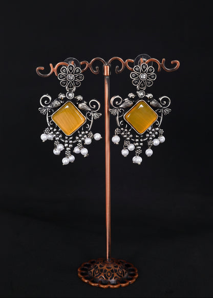 A pair of ornate silver-toned statement earrings with large square amber-yellow centers and dangling white pearl-like beads.