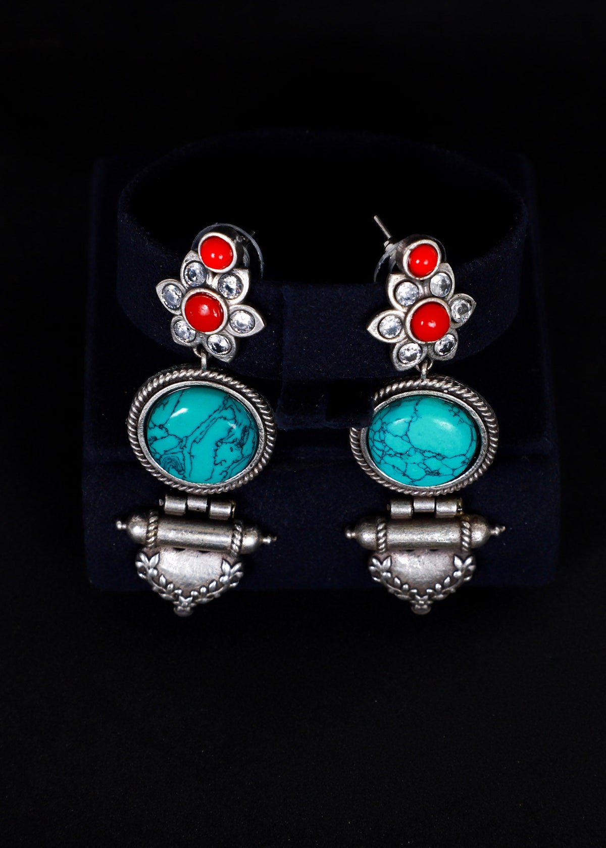 A pair of silver-toned dangle earrings. Each earring features a silver floral stud with three round orange-red stones, a round turquoise-blue stone in a silver bezel, and a dangling silver bell-shaped charm.