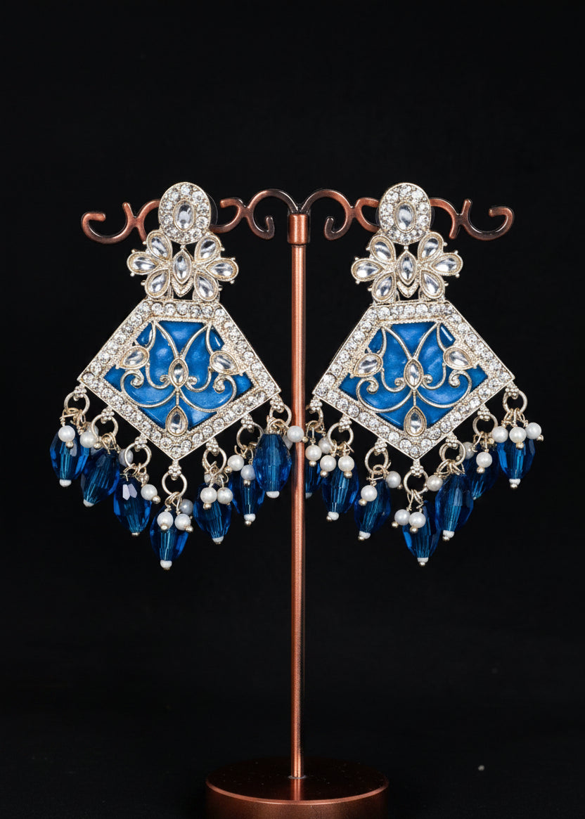 A pair of ornate blue and silver chandelier earrings. The earrings feature a silver-toned filigree frame set with clear rhinestones, a vibrant blue enamel center, and dangling blue crystal beads and pearl accents.