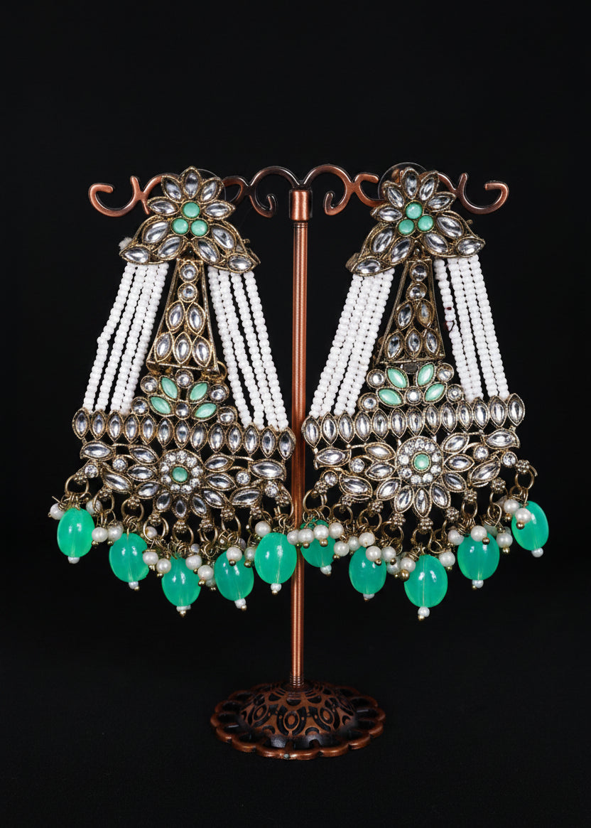 A pair of ornate Indian chandbali earrings. The earrings feature silver-toned metalwork, white bead strands, and green stone accents.