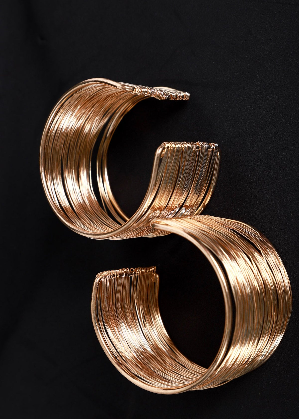 Two wide metallic cuff bracelets with a stacked, layered look in a warm gold tone, displayed on a solid black background.