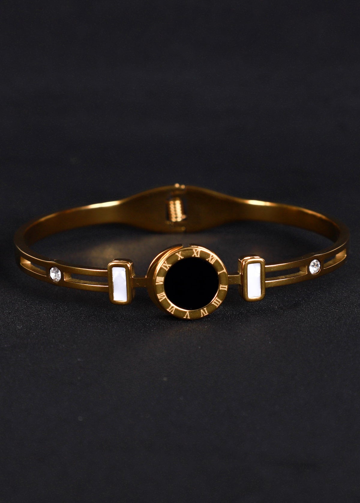 A gold-plated bangle bracelet with a central black circular bezel, two white rectangular inlays, and two small clear stones, shown on a dark background.