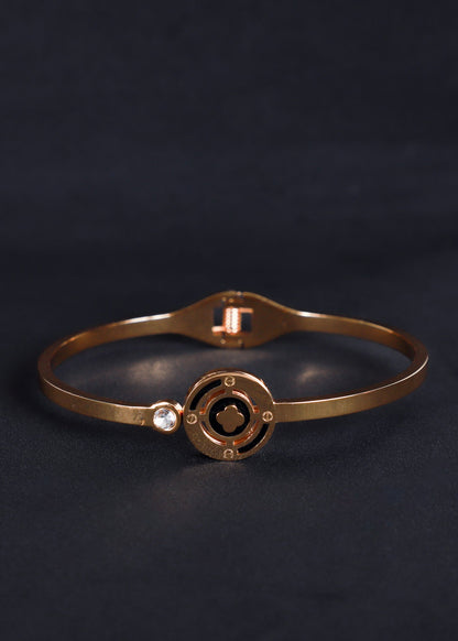 A slim rose gold bangle bracelet with a circular medallion design and a small clear stone accent, displayed on a dark textured background.