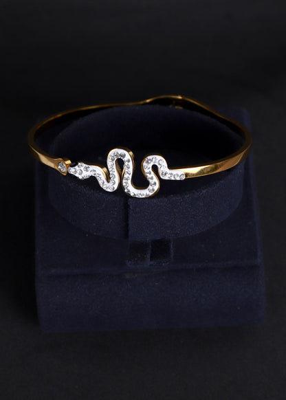 A gold snake-shaped cuff bracelet adorned with clear rhinestones, displayed on a dark navy velvet jewelry box against a black background.