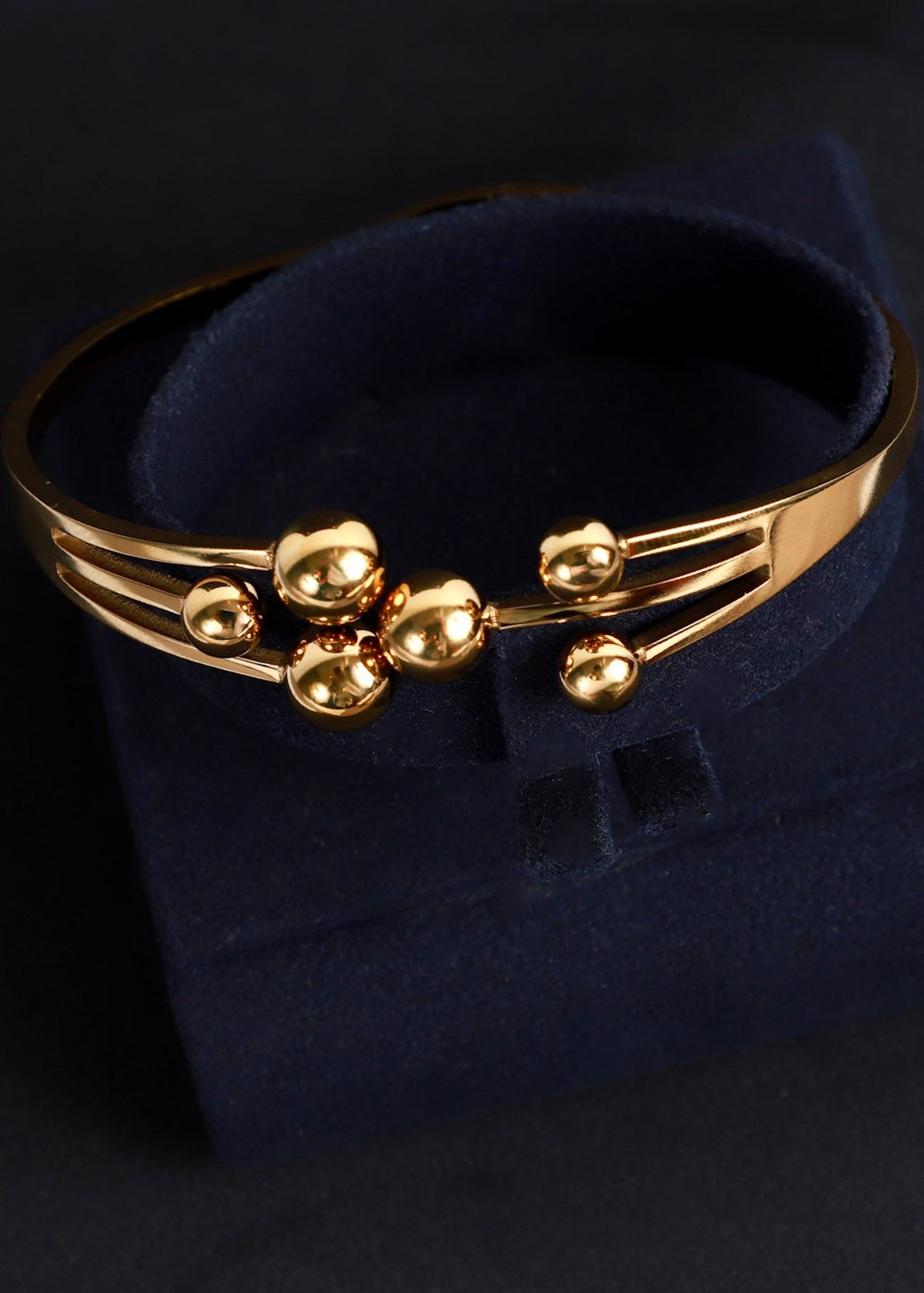 A gold-toned metal cuff bracelet with three raised oval bead-like accents on the front, displayed on a dark navy velvet jewelry box against a black background.