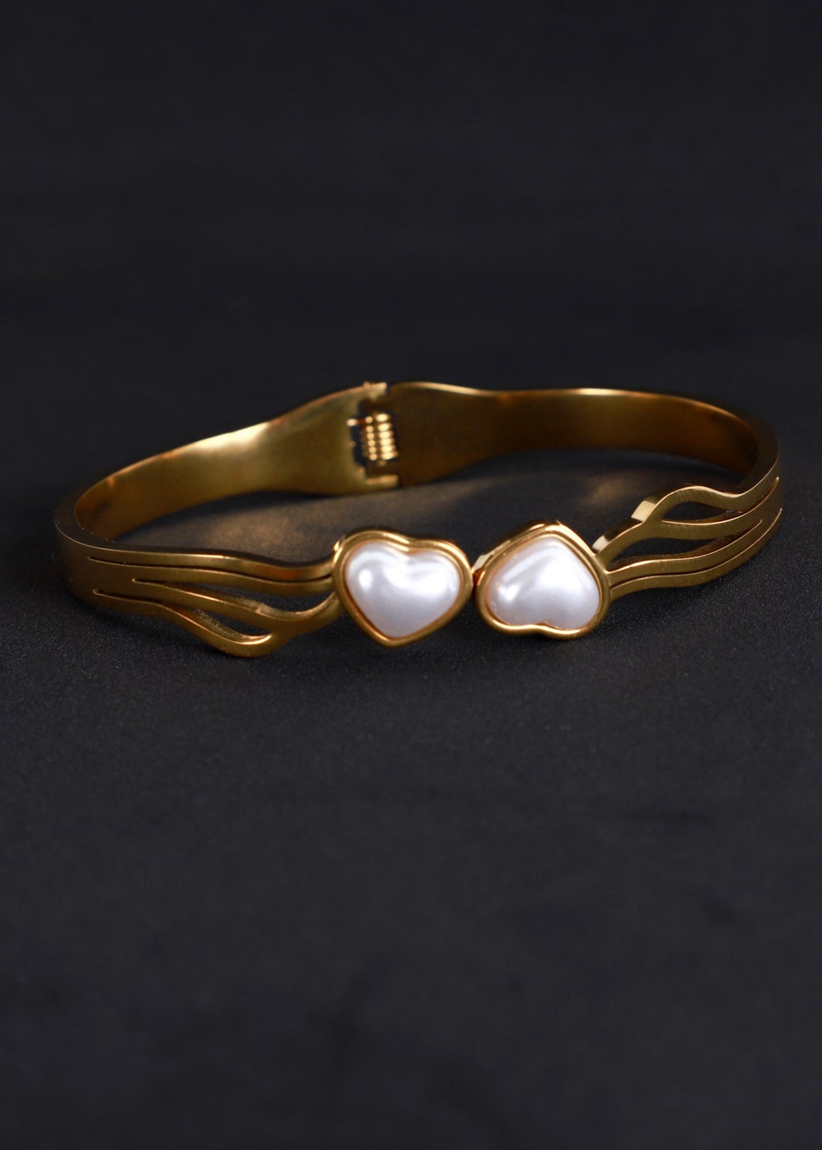 A gold-plated metal cuff bracelet featuring two heart-shaped white mother-of-pearl inlays, displayed on a dark textured background.