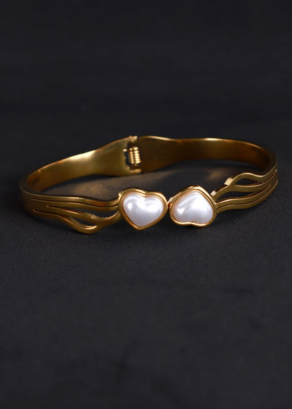 A gold-plated metal cuff bracelet featuring two heart-shaped white mother-of-pearl inlays, displayed on a dark textured background.
