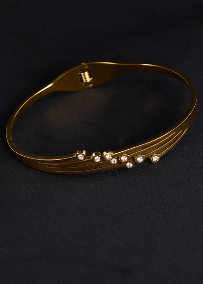A gold-toned metal bangle bracelet with a row of small clear rhinestones, displayed on a dark textured background.