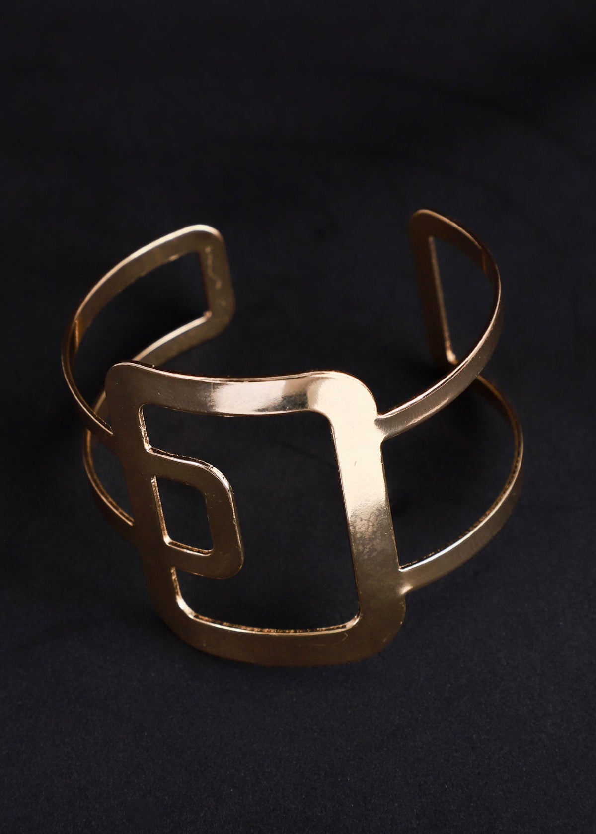 A wide, open cuff bracelet in a brushed gold finish, featuring a bold, geometric square frame design, displayed on a dark textured background.