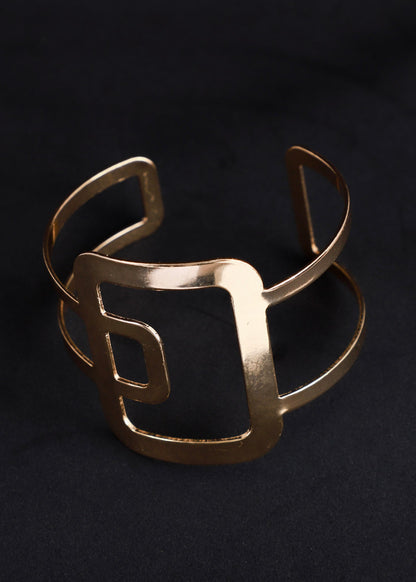 A wide, open cuff bracelet in a brushed gold finish, featuring a bold, geometric square frame design, displayed on a dark textured background.