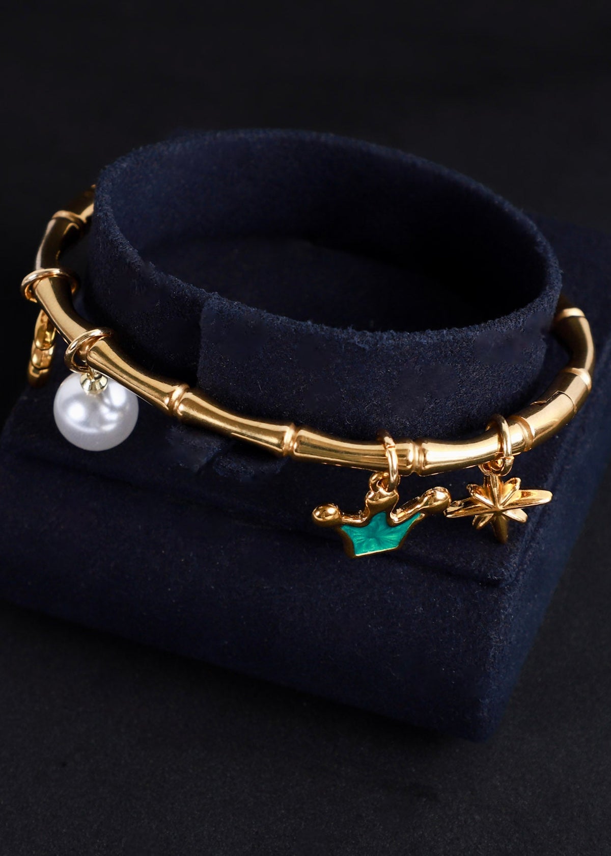 A gold-toned charm bracelet displayed on a dark navy velvet jewelry stand against a black background. The bracelet features a bamboo-inspired segmented band with dangling charms including a white pearl, a green leaf, and gold-toned floral/star details.