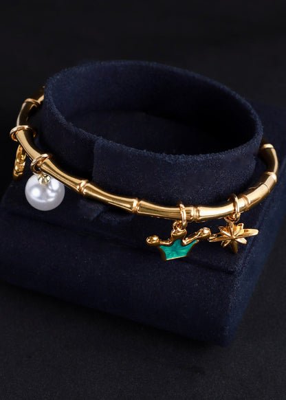 A gold-toned charm bracelet displayed on a dark navy velvet jewelry stand against a black background. The bracelet features a bamboo-inspired segmented band with dangling charms including a white pearl, a green leaf, and gold-toned floral/star details.