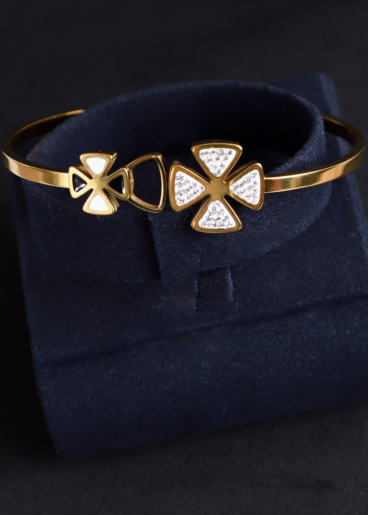 A gold-plated bangle bracelet with three decorative cross-like motifs, including one adorned with small clear stones, displayed on a dark navy velvet jewelry box against a dark background.