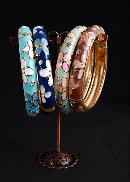 Four decorative bangles displayed on a small stand against a black background. The bangles have floral patterns in light blue, navy blue, mint green, and brown, with gold-toned metal outlines and rhinestone accents.