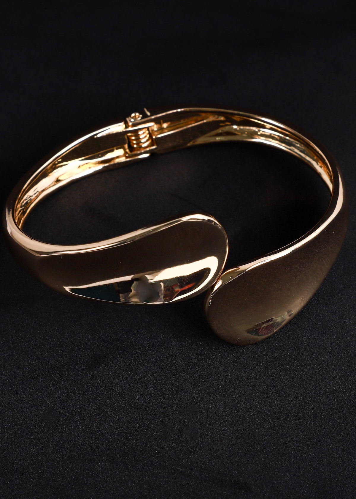 A polished gold-toned metal cuff bracelet with a smooth, curved organic design, displayed on a dark textured background.
