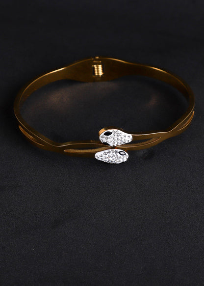 A gold-plated bangle bracelet with two small fish charms encrusted with clear rhinestones, displayed on a black textured background.