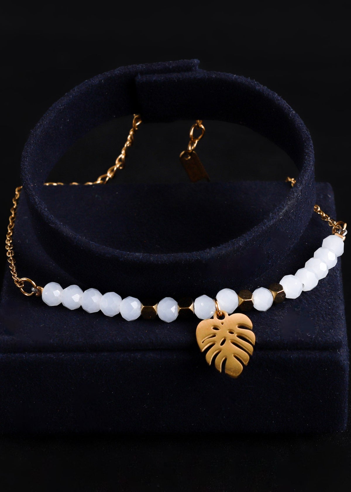 A gold-tone beaded necklace with white pearl-like beads and a gold monstera leaf pendant, displayed on a black velvet jewelry stand against a black background.