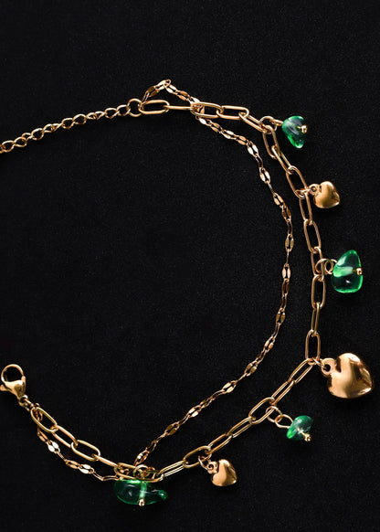 A gold-toned multi-strand chain bracelet with dangling heart and green bead charms, displayed on a solid black background.