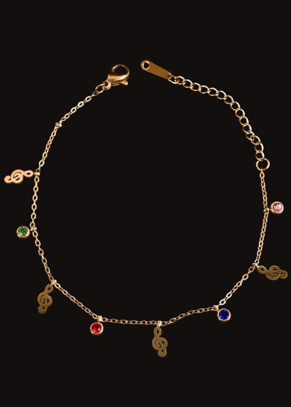 A delicate gold chain bracelet with musical note charms and small colorful gemstone accents, displayed on a black background.
