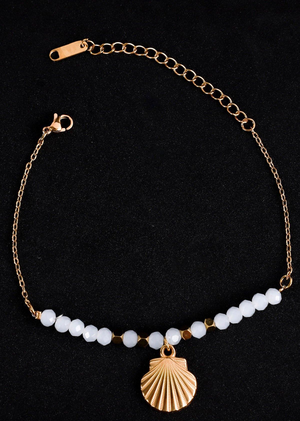 A gold chain anklet featuring a row of small white beads and a gold shell-shaped charm, displayed on a black background.