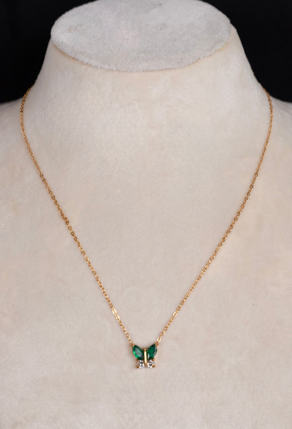 A delicate gold chain necklace with a butterfly pendant featuring green wings and small clear stones, displayed on a cream-colored jewelry bust.