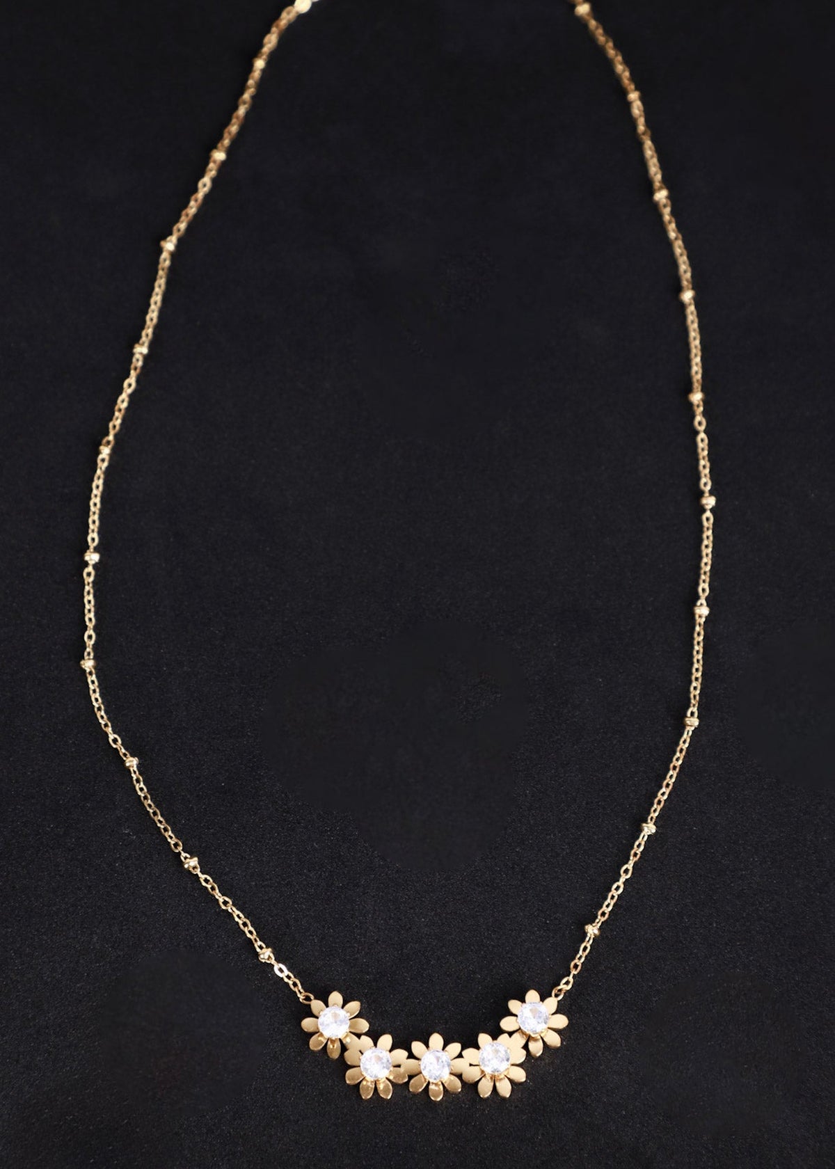 A delicate gold chain necklace featuring a row of small flower charms with white centers, displayed against a dark black background.