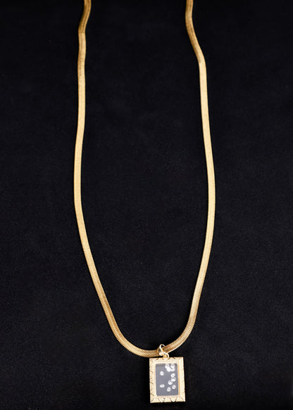 A gold snake chain necklace with a rectangular pendant featuring a gray and white starry night sky design, displayed against a black background.
