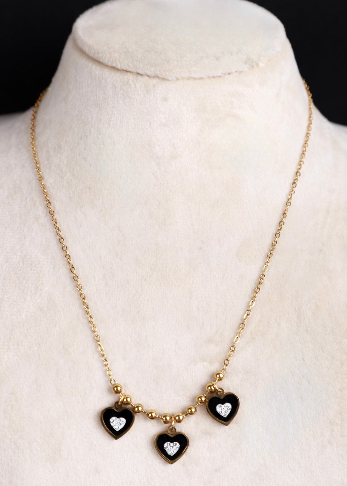 A gold chain necklace displayed on a cream-colored jewelry bust, featuring three small heart charms with black enamel and clear heart-shaped stones.