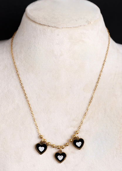 A gold chain necklace displayed on a cream-colored jewelry bust, featuring three small heart charms with black enamel and clear heart-shaped stones.