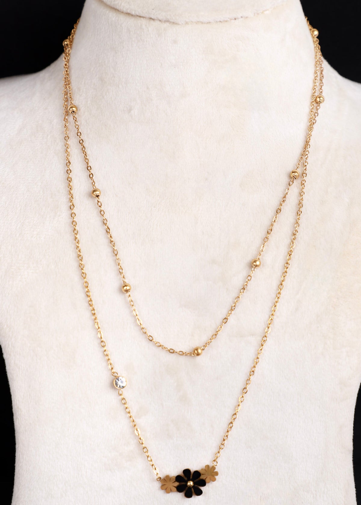 A gold-tone double-layered necklace displayed on a white mannequin bust. The necklace features a delicate chain with small gold beads and a lower pendant layer adorned with a black flower and gold leaf accents.