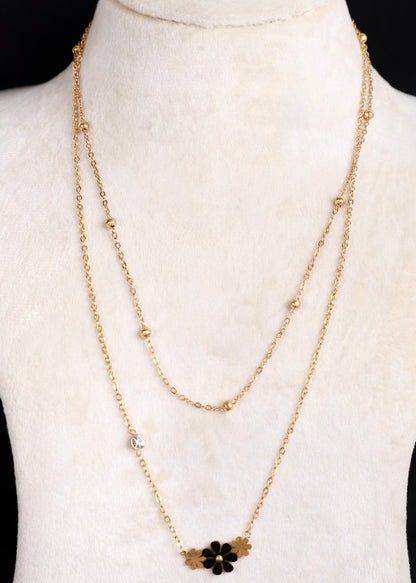 A gold-tone double-layered necklace displayed on a white mannequin bust. The necklace features a delicate chain with small gold beads and a lower pendant layer adorned with a black flower and gold leaf accents.