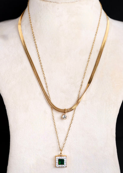 Anti tarnish gold layered necklace set displayed on a cream-colored jewelry bust. The set features a wide snake chain, a delicate cable chain, a small drop pendant, and a longer chain with a square pendant set with a green gemstone and clear accent stones.