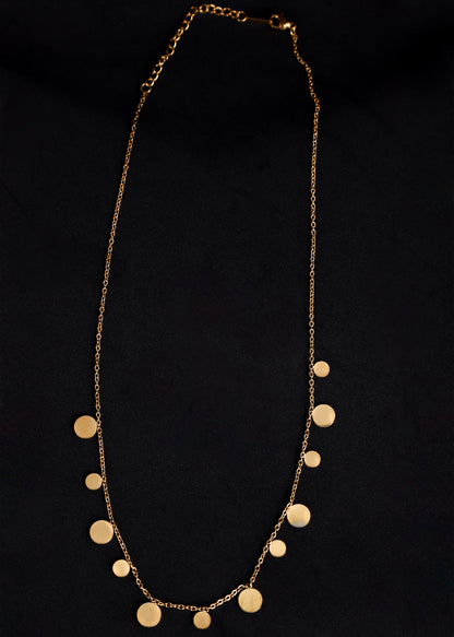 A gold chain necklace featuring multiple flat, disc-shaped metal pendants of varying sizes, displayed against a solid black background.