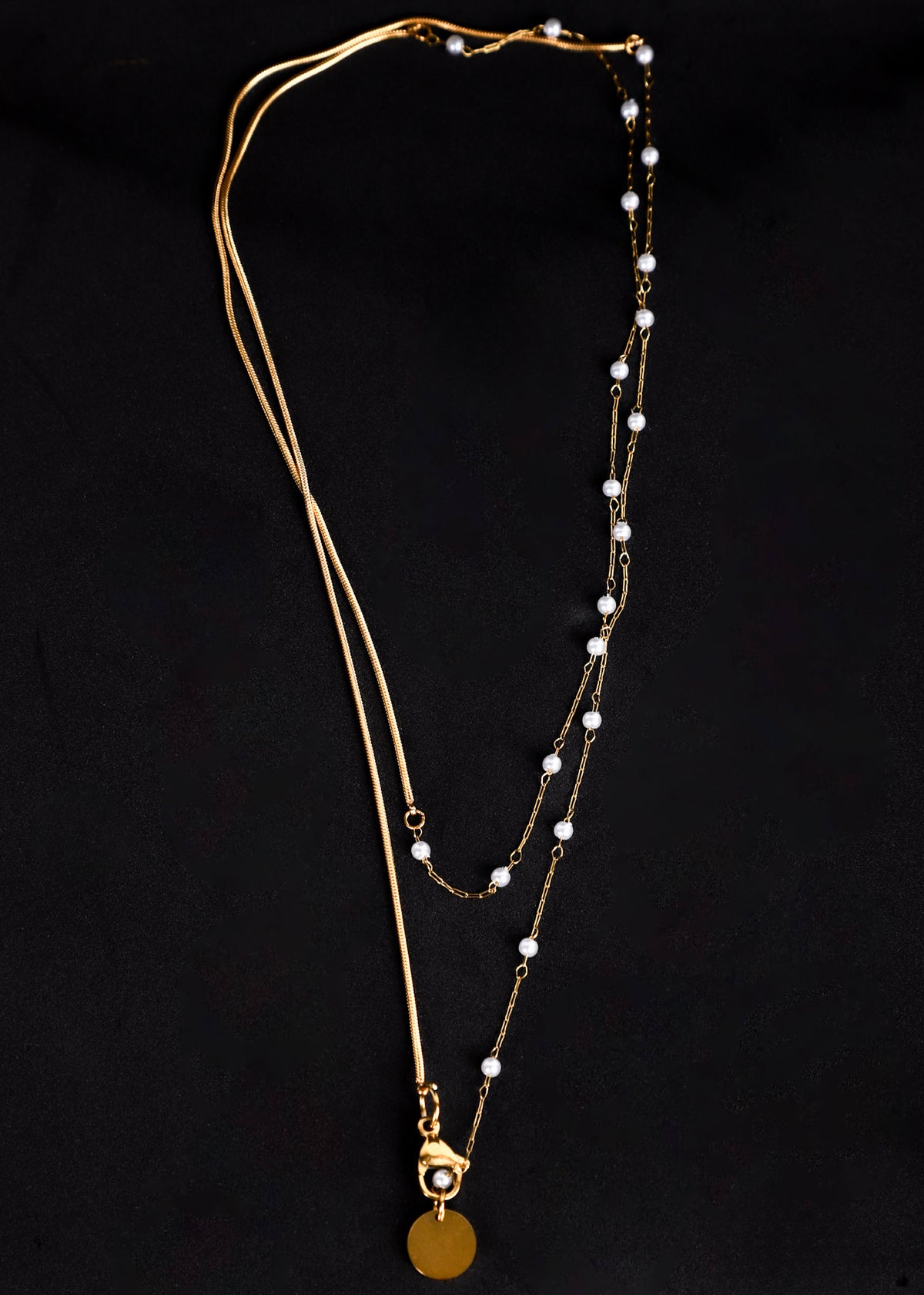 A gold-toned double-layered necklace featuring a delicate chain with small white pearl beads and a round gold disc pendant, displayed against a solid black background.