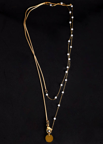 A gold-toned double-layered necklace featuring a delicate chain with small white pearl beads and a round gold disc pendant, displayed against a solid black background.