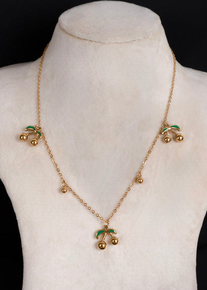 A gold-tone cherry necklace displayed on a cream-colored mannequin bust against a black background. The necklace features three clusters of cherries with green enamel leaves and small gold beads along the chain.