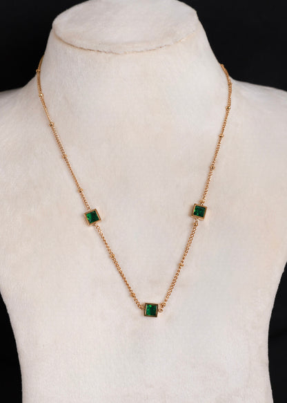 A gold anti-tarnish chain necklace with three small square green gemstones, displayed on a cream-colored jewelry bust against a black background.