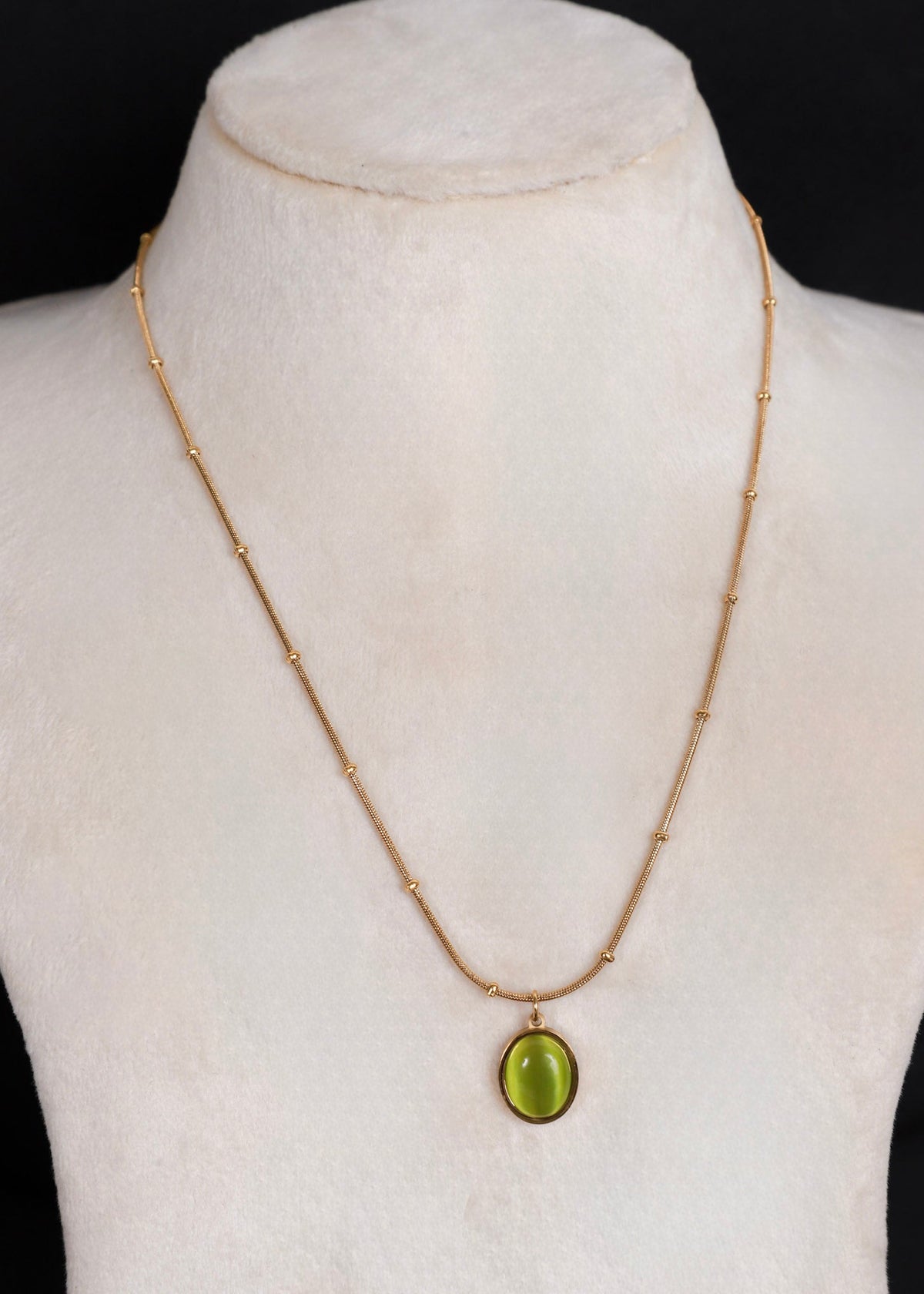 A gold chain necklace with a small oval green pendant, displayed on a cream-colored mannequin bust against a black background.