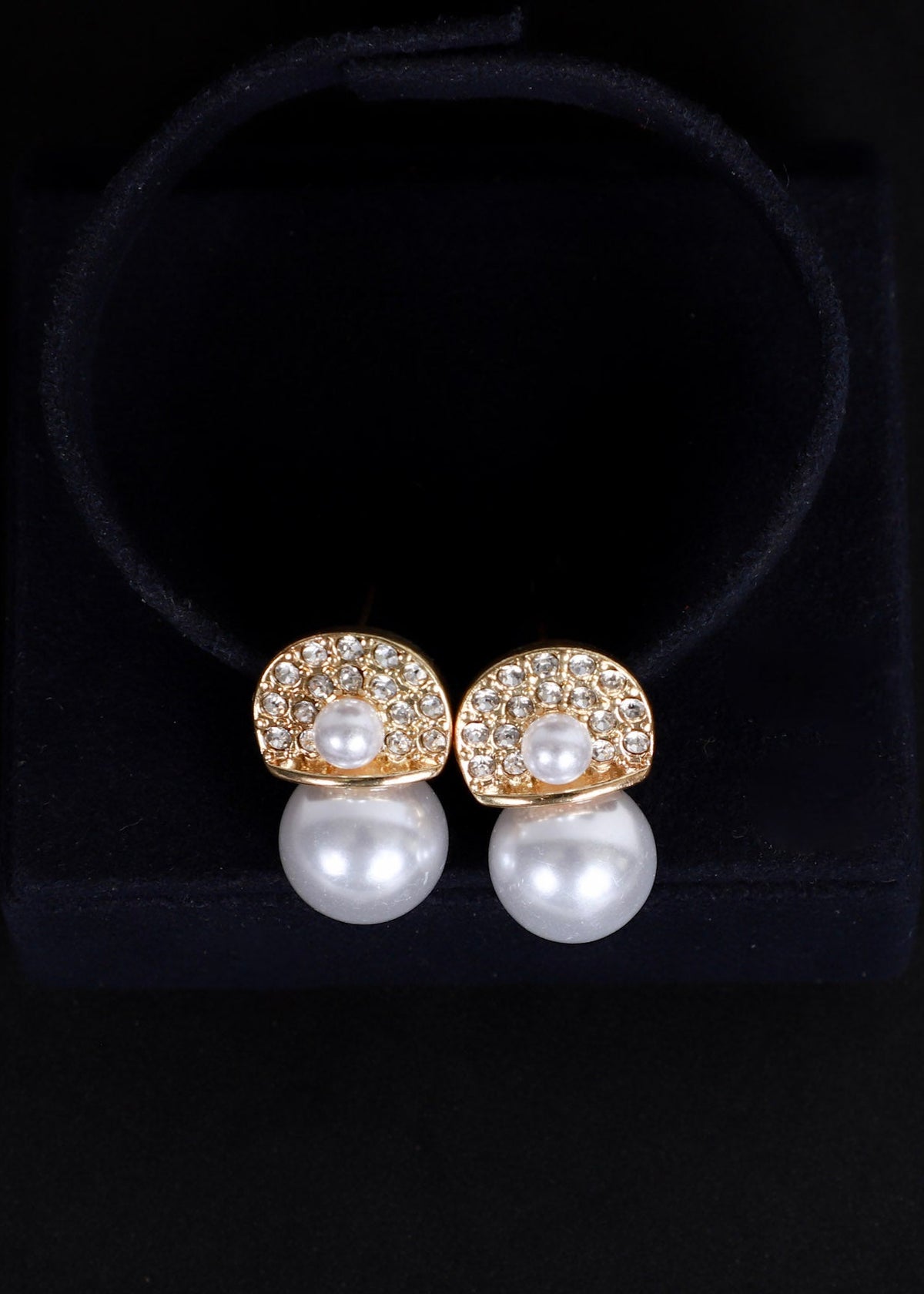 A pair of gold and pearl stud earrings displayed on a black velvet jewelry stand against a black background.