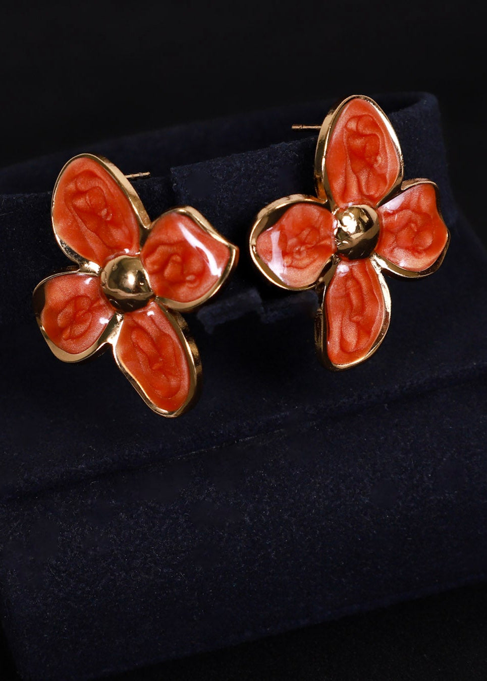 A pair of gold-tone flower-shaped cufflinks with orange-red enamel petals, displayed on a dark navy velvet jewelry stand against a black background.