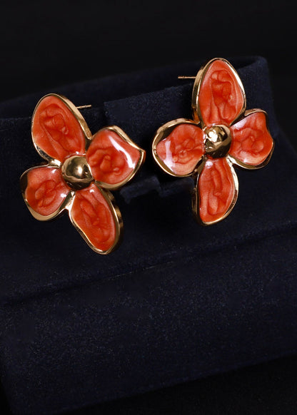 A pair of gold-tone flower-shaped cufflinks with orange-red enamel petals, displayed on a dark navy velvet jewelry stand against a black background.