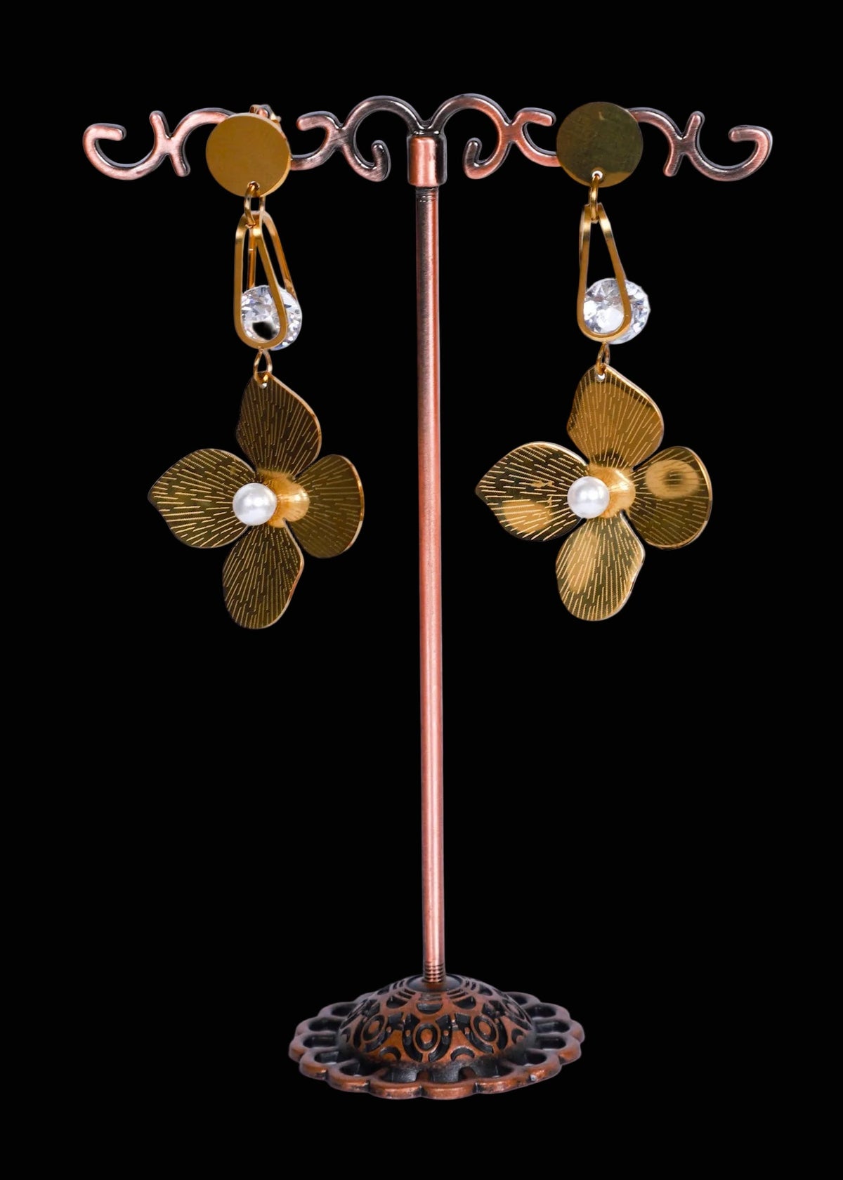 A pair of dangling flower earrings. The earrings have textured bronze-toned petals with a small white pearl-like center and clear hanging stones.