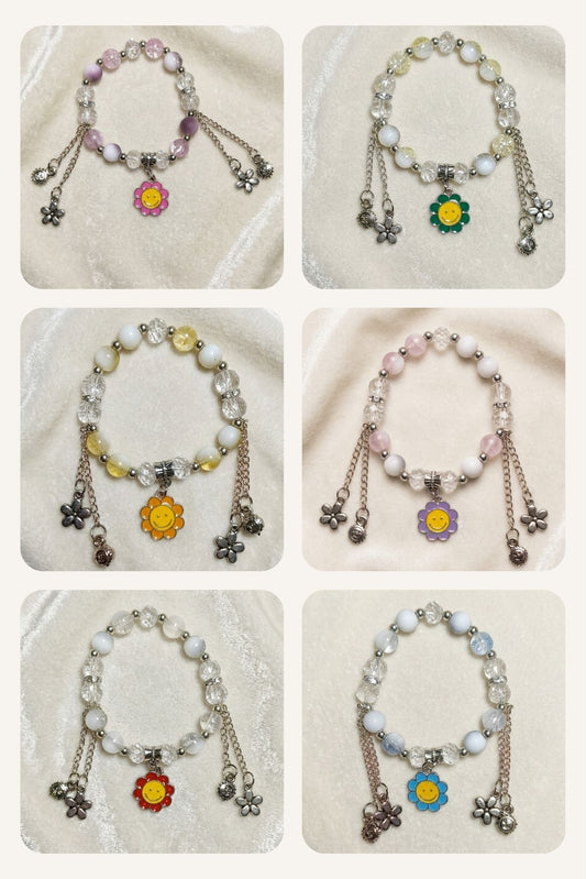 Six beaded charm bracelets arranged in a 3x2 grid on a soft cream fabric background. Each bracelet features white pearl-like beads, silver-toned chains, dangling flower charms, and small star charms.